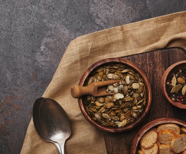 a bowl of pumpkin seeds on a table with a spoon and linens, with some small bread next to it. health benefits of pumpkin seeds, roasted pumpkin seeds, benefits of pumpkin seeds, nutrition benefits of pumpkin seeds, health benefits of pumpkin seed, health benefits of pepitas, pepitas, roasted pepitas