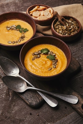 bowls of pumpkin soup with pumpkin seed garnish on top. Pumpkin seeds in a bowl, with a spoon, and bread, and large spoons next to it. health benefits of pumpkin seeds, roasted pumpkin seeds, benefits of pumpkin seeds, nutrition benefits of pumpkin seeds, health benefits of pumpkin seed